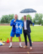 Two men on a rainy soccer field pose with a blue KANGASLAHTI 44 jersey, one holding an umbrella and smiling excitedly