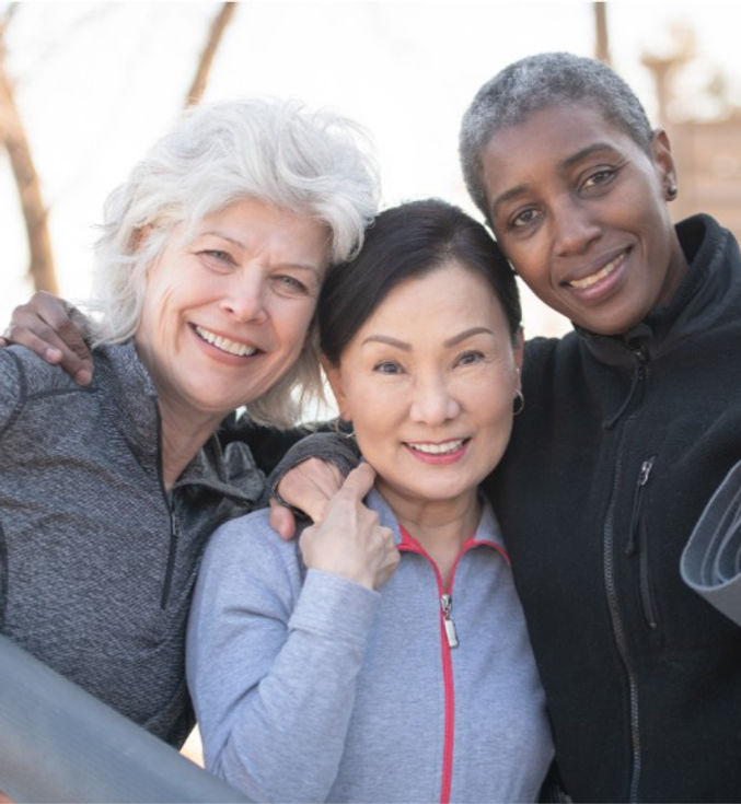 3 female older adults close up with arms around each other while smiling and holding yoga mats