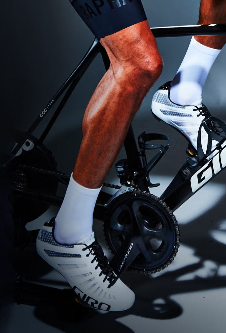 Cyclist on a black bike, pedaling in white shoes and socks. Dark background with visible leg muscles and "GIRO" text on shoes.