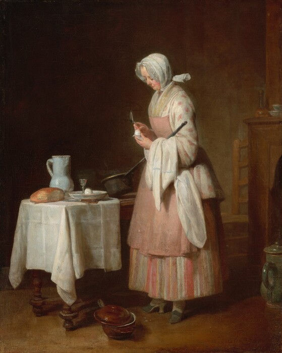 A woman holds an egg and spoon beside a table set with bread and kitchen vessels.