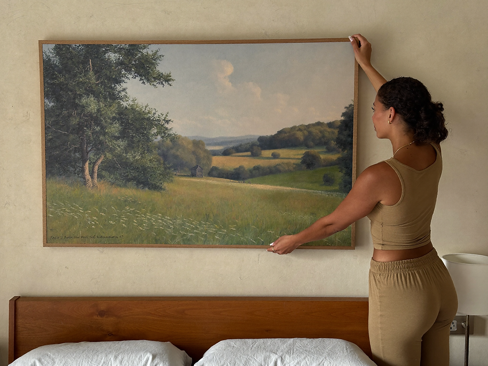 Woman gently adjusting large meadow wall art above a linen bed in a calm neutral bedroom.