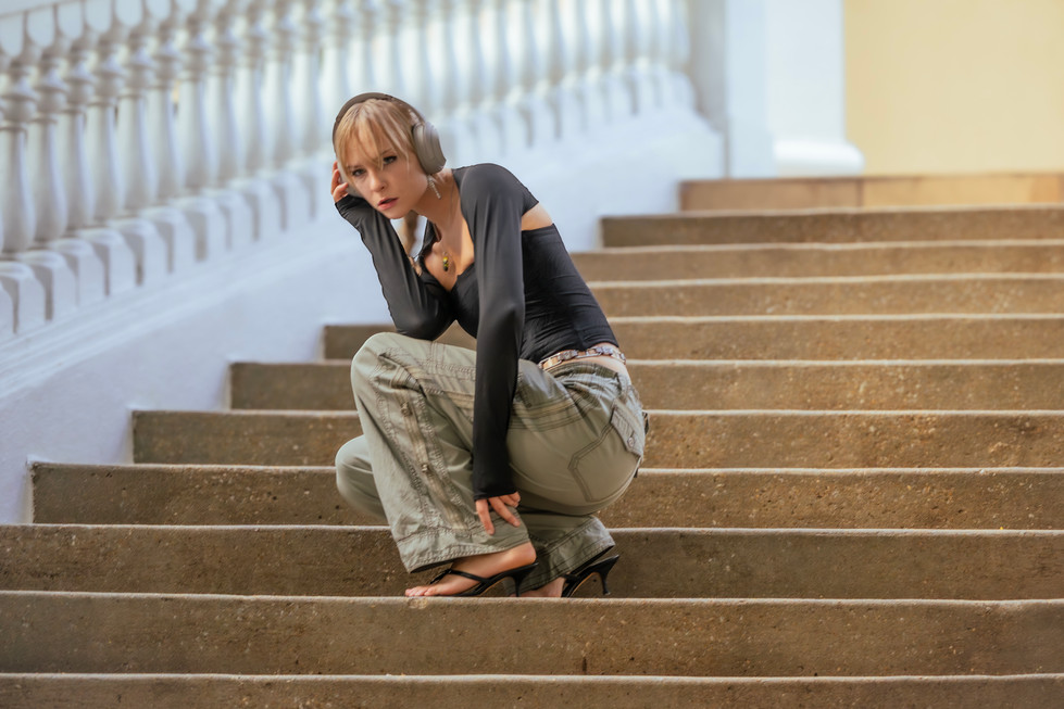 White woman crouching on the steps for a pose. She leans to one side while her hands grabs one ear