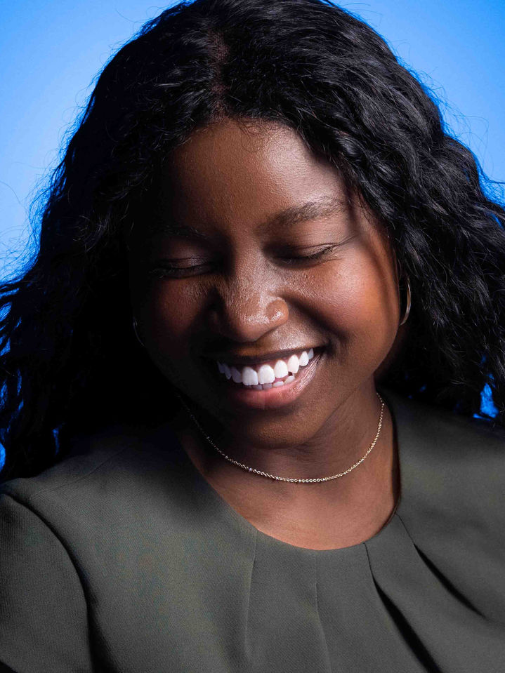 Portrait studio d’une femme noire souriante au regard baissé, sur fond bleu éclatant, photographiée par Frédéric Pace.