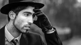 Portrait dramatique d’un comédien en costume, chapeau Borsalino et gant de cuir, regard inquiet, la main sur le front, incarnant un personnage troublé dans une ambiance de polar.