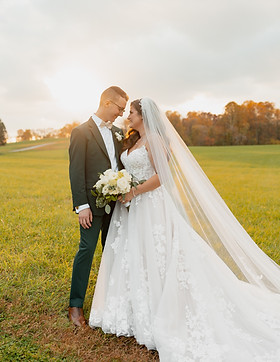 A bride and groom stand on a grassy field at sunset, captured by a talented Lancaster wedding photographer. The bride wears a long white gown and holds a bouquet of white flowers, while the groom is in a dark suit. They gaze affectionately at each other under the cloudy sky.