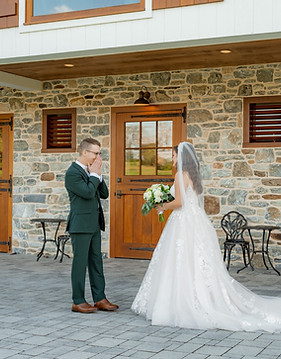 A bride and groom stand facing each other outside a rustic building with stone walls and wooden doors. The bride wears a white gown, and the groom is in a dark suit. The scene suggests a wedding ceremony or photo shoot.