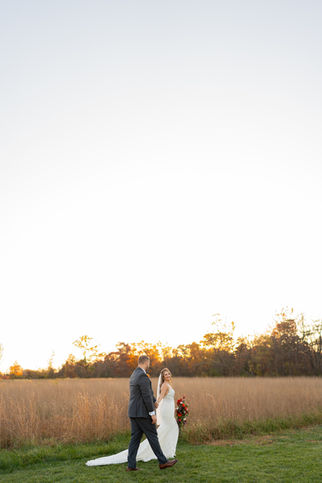A bride and groom walk hand in hand in a grassy field during sunset, captured beautifully by a PA wedding photographer. The bride wears a long white gown with a veil, carrying a bouquet of red flowers, while the groom dons a dark suit against autumn trees and a clear sky.