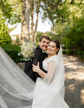 A bride and groom stand close together, embracing under a natural canopy of green leaves. The bride is in a white dress, and the groom is in a black suit. Both are smiling, conveying a joyful and romantic moment.