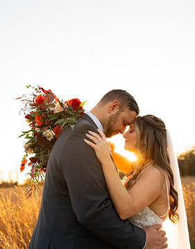 A couple embraces tenderly in an outdoor setting, with a blurred bouquet of red and green flowers in the background. The woman wears a yellow top, and the atmosphere is warm and romantic.