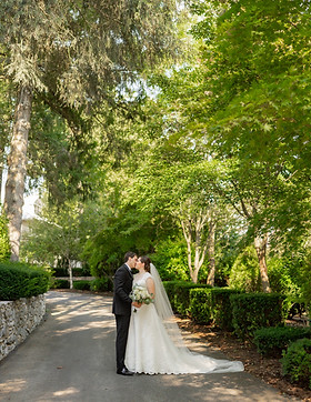 A couple in wedding attire shares a kiss on a tree-lined pathway, elegantly captured by a Lancaster wedding videographer. The brides flowing white dress and veil complement the grooms black suit, as lush greenery surrounds them, creating a romantic and serene backdrop.