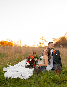A bride in a white gown with a long train and a groom in a gray suit sit on a grassy field during sunset, captured beautifully by a Central PA wedding photographer. The bride holds a bouquet of red and orange flowers, while trees in the background reflect the golden hour light.