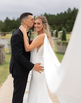 A groom in a black suit kisses a smiling bride on the forehead. The bride, wearing a white dress with a cape, stands on a stone path with a green, tree-lined background. They embrace, and the brides veil is flowing.
