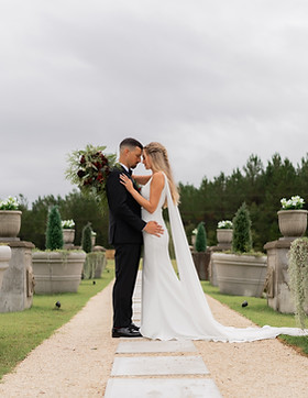 A bride and groom stand closely on a garden path, captured beautifully by a Lancaster wedding photographer. The bride, in a long white dress with train and veil, holds a bouquet. The groom wears a black suit. They touch foreheads amid greenery and large planters under a cloudy sky.