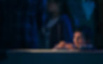 A young boy leans on a barrier under warm light, looking down thoughtfully, while people stand behind him in a dimly lit indoor setting.