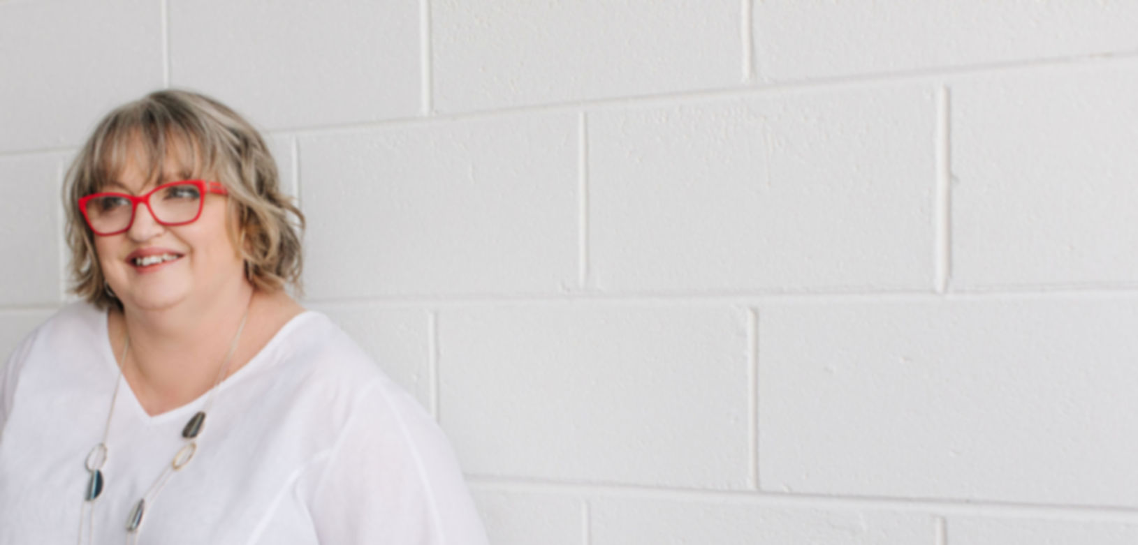 Di Geddes standing in a white top and red glasses, smiling in front of a white brick wall