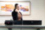 Person with headset talking behind a counter. JBL speaker and HP monitors visible. Blurred colorful art in the background. Office setting.