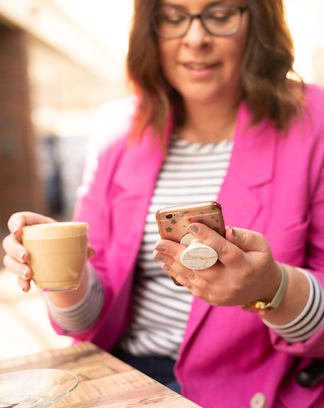 Laura is looking at her phone and holding a cup of coffee, she is wearing a pink blazer