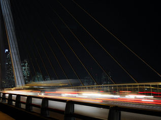 Night view of a cable-stayed bridge with red and white car light trails and glowing city skyscrapers in the background