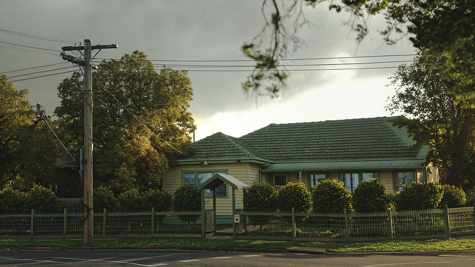 Suburban house with green roof and fenced yard under power lines, lit by sun breaking through cloudy sky