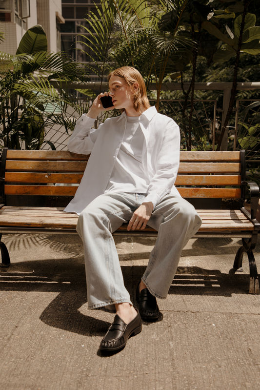 erson seated on a wooden park bench, on a call, wearing the Atelier N22 Edition 01 white linen shirt open over a white tee, light-wash wide-leg jeans, and black loafers. Urban greenery and building facade in background. Hong Kong.