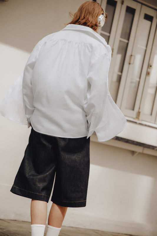 Back view of a person in motion, wearing the Atelier N22 Edition 01 white linen shirt with dark denim shorts and white socks. Shirt fabric catching wind, relaxed movement. Courtyard setting.