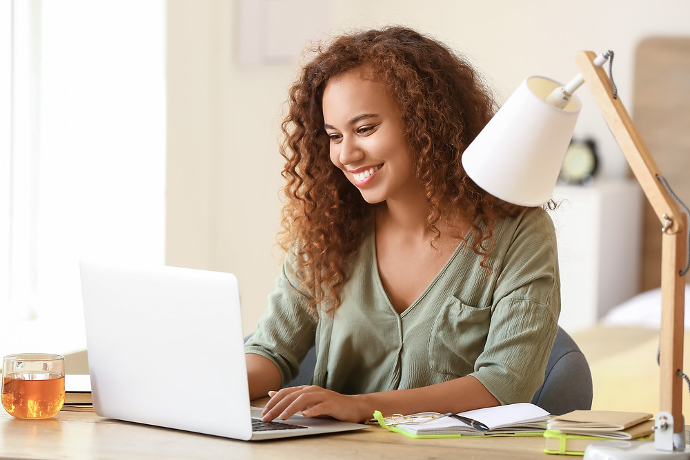 A smiling student sits at a desk with an open laptop and with notebooks