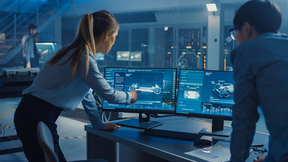 Two business people stand over a desktop screen with diagrams of a car design on.