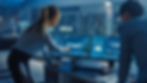 Two business people stand over a desktop screen with diagrams of a car design on.