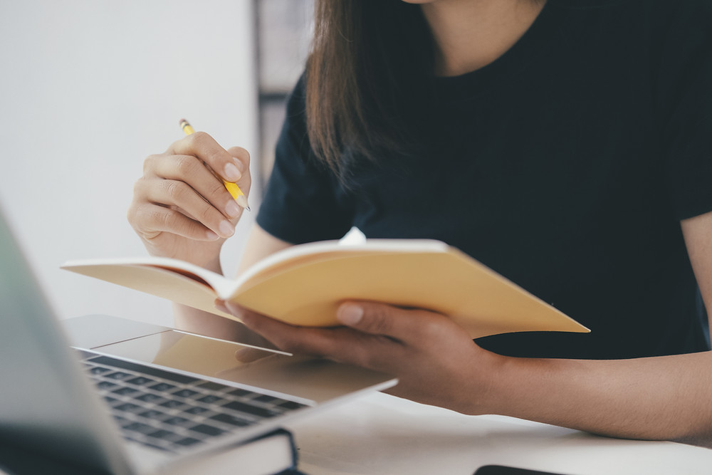 A student is studying at their desk in front of a laptop and is holding an open notebook and a pencil