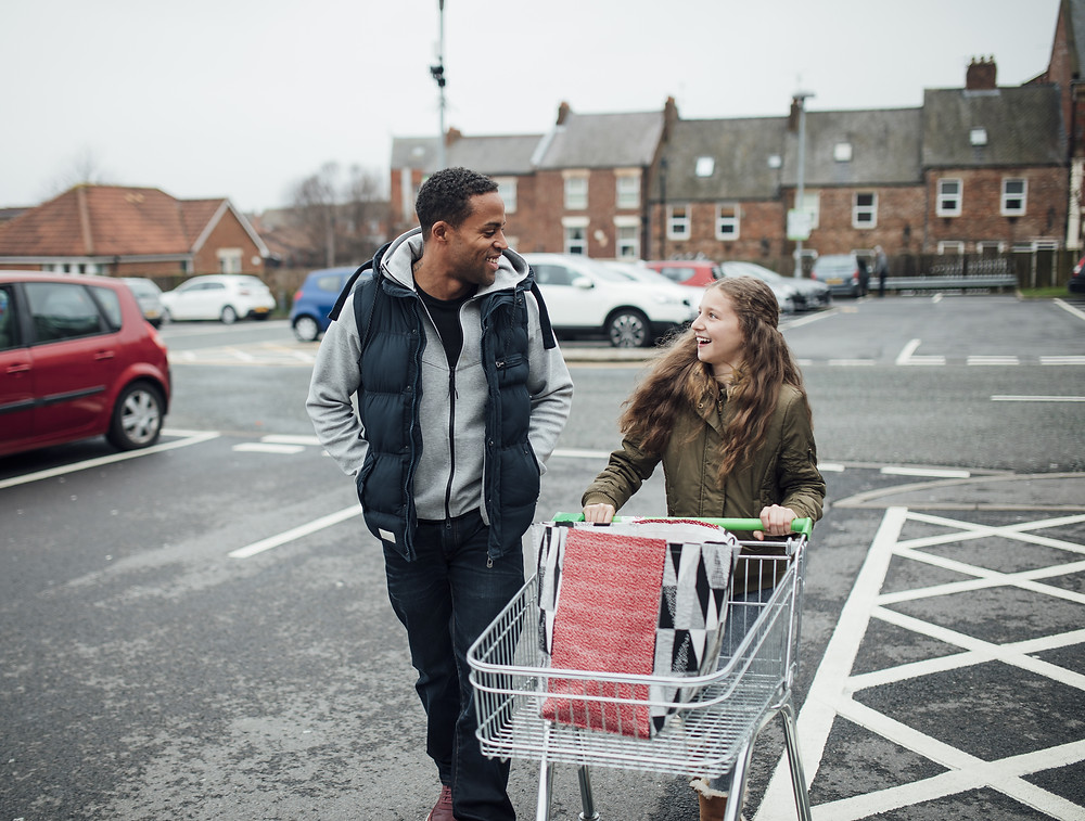 Two young adults are laughing together. They are walking across a supermarket carpark. One of them is pushing a trolley with a carrier bag in.
