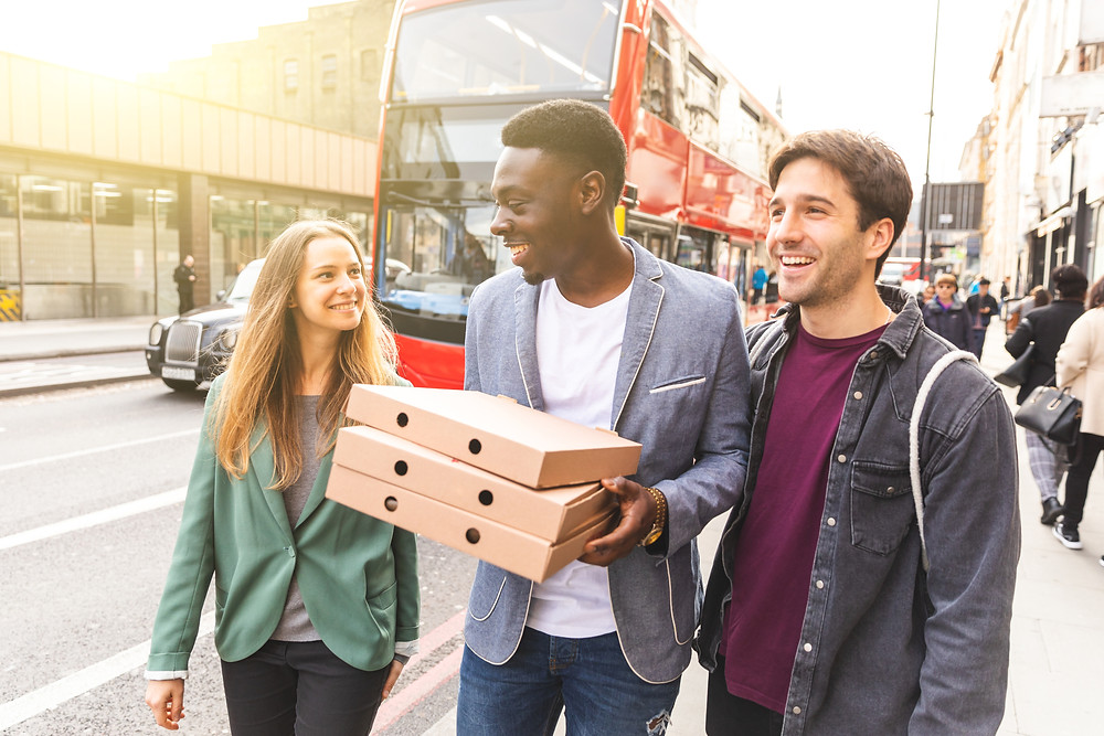 Three young adults walk down a busy street in London and are laughing together. The person in the middle is carrying three pizza boxes. In the background there are other pedestrians and a red double-decker bus and a black cab.