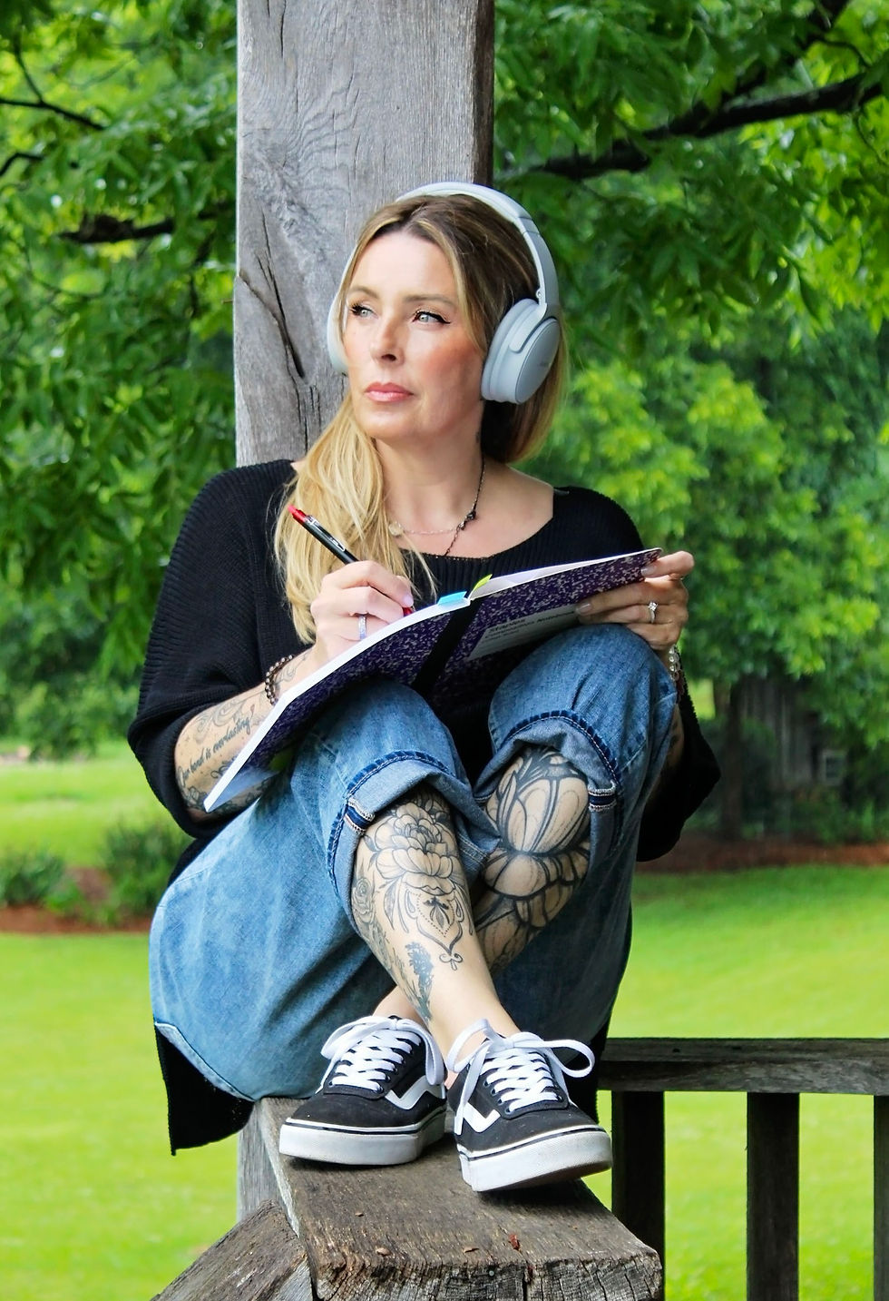 Woman with tattoos writing in notebook outdoors with headphones