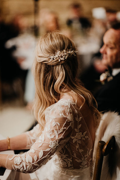 The bride, wearing an intricately designed white lace gown, rests her hand on the table as she sits at a reception, her blonde hair adorned with a delicate, jeweled hairpiece.