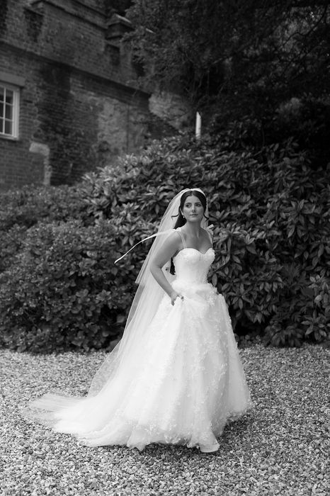 A bride in long, flowing white dress and veil walks toward the camera in front of a stone building and dense shrubbery.