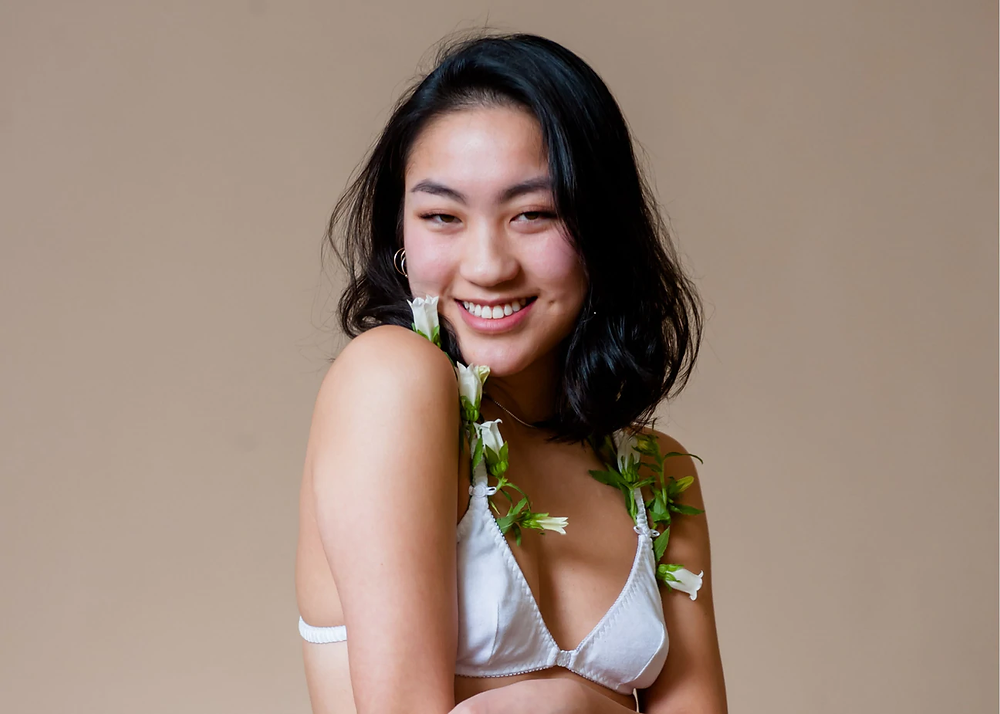 Smiling women with short hair, natural makeup, and wearing a bralette. The bralette has white flowers poking out, adds to the aesthetic of femininity and empowerment.
