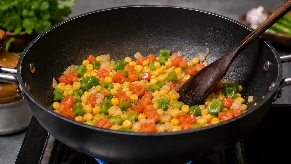 Colorful corn, peas, and diced tomatoes cooking in a black pan with a wooden spatula. Herbs and ingredients blurred in the background.