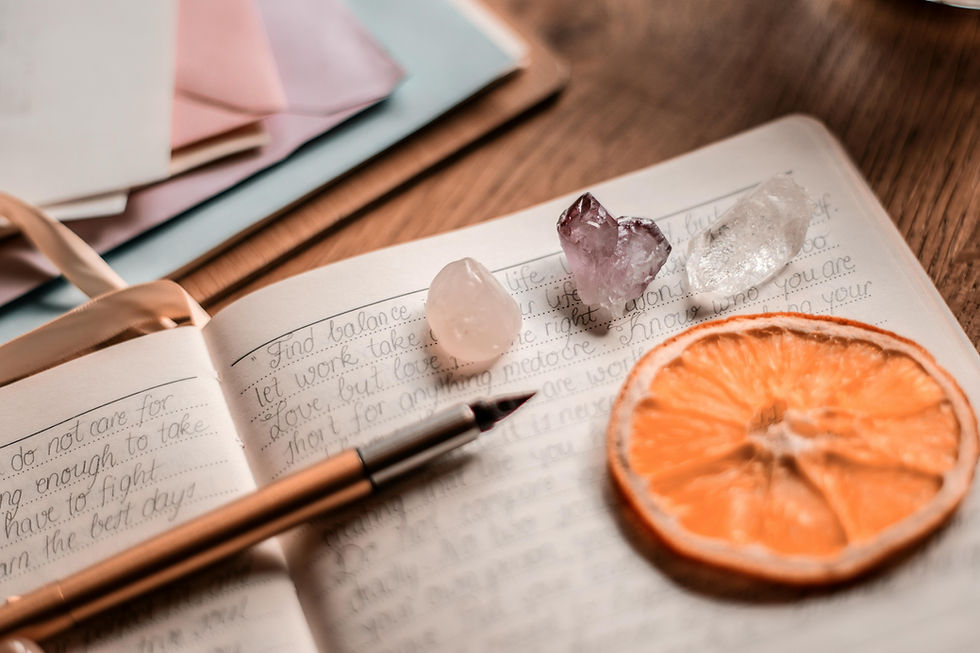 Open journal with handwritten notes, a pen, and three crystals (rose quartz, amethyst, and clear quartz) placed on the page beside a dried orange slice on a wooden table, creating a calm and mindful journaling scene.