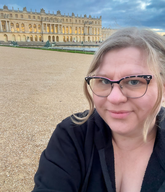 A blond middle aged woman takes a selfie in front of the Palace of Versailles.