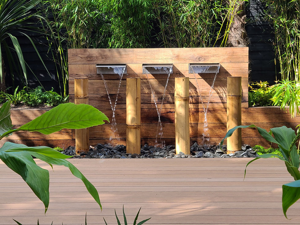 Three bamboo fountains pour water over pebbles in a lush garden with greenery. A wooden deck is in the foreground, creating a peaceful scene within a tropical garden in Chorlton, designed by DK Garden Design.