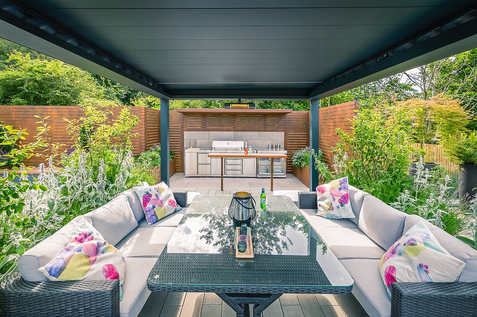 Modern patio with a glass table and colourful cushions on a sofa, and an outdoor kitchen and greenery in background, designed by David Keegan Garden Design.