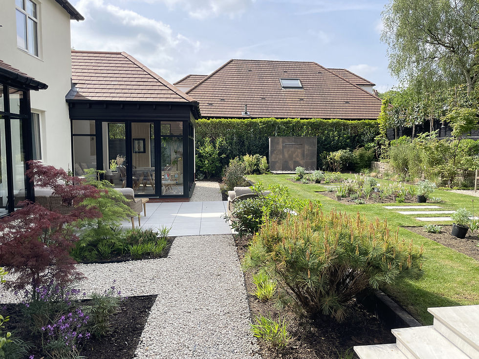 A modern garden in Marple Bridge designed by David Keegan from DK Garden Design, with a gravel path, patio and lush greenery. A house with a glass extension overlooks the landscaped lawn under a sunny sky.