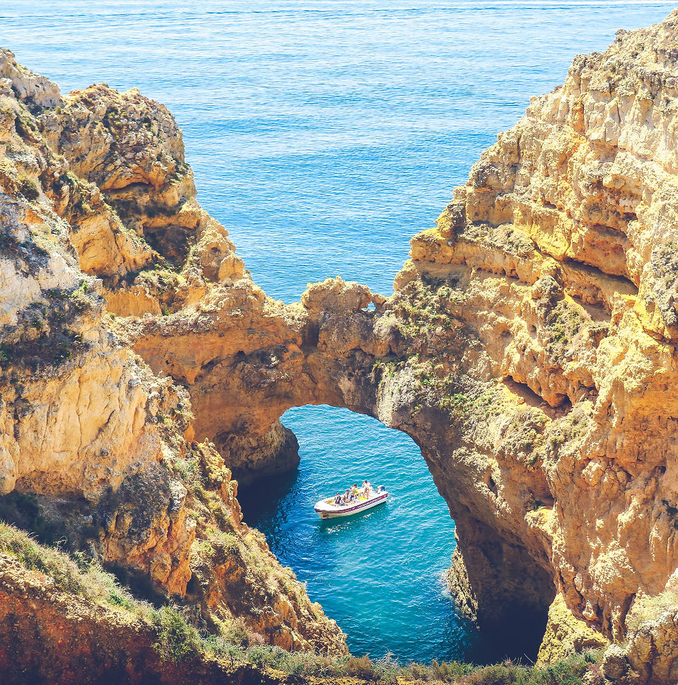 Tour boat passes under a rocky sea arch between golden cliffs, with bright blue water below and open ocean beyond.