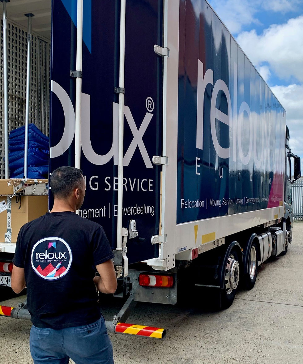 Man in a Reloux shirt stands beside a branded moving truck being loaded, with boxes and blue blankets visible under a cloudy sky.