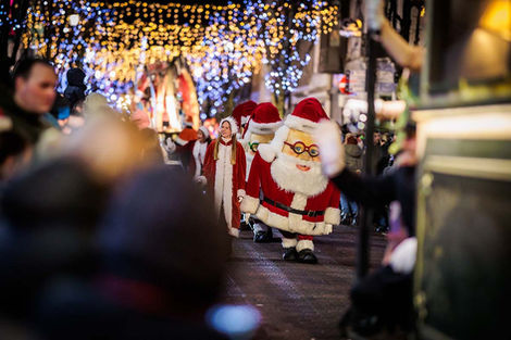 Kerstparade met een lopende kerstmanmascotte onder feestverlichting.