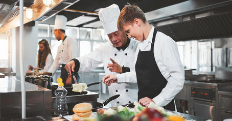 Chefs in a kitchen, with one guiding another on cooking. Both wear white uniforms and hats. Bright setting with pans, veggies, and bread.