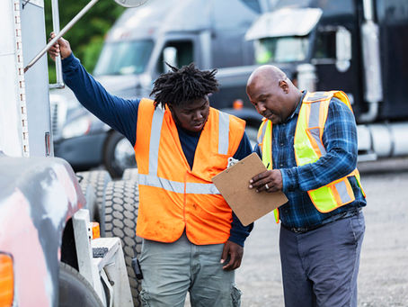 Two workers in orange safety vests review a clipboard outdoors near parked trucks. One points to the truck, focused and attentive.