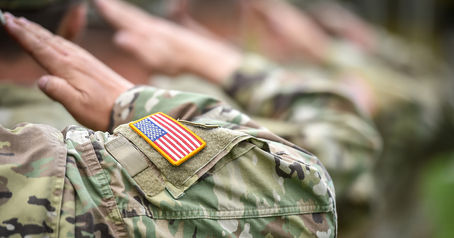 Soldiers in camo uniforms saluting, with U.S. flag patch visible on one sleeve. The background is blurred, creating a focused, respectful scene.