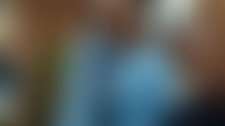 Woman in a blue dress leans over a man with glasses, focusing on work. Brick wall and plants in office background. Serious mood.