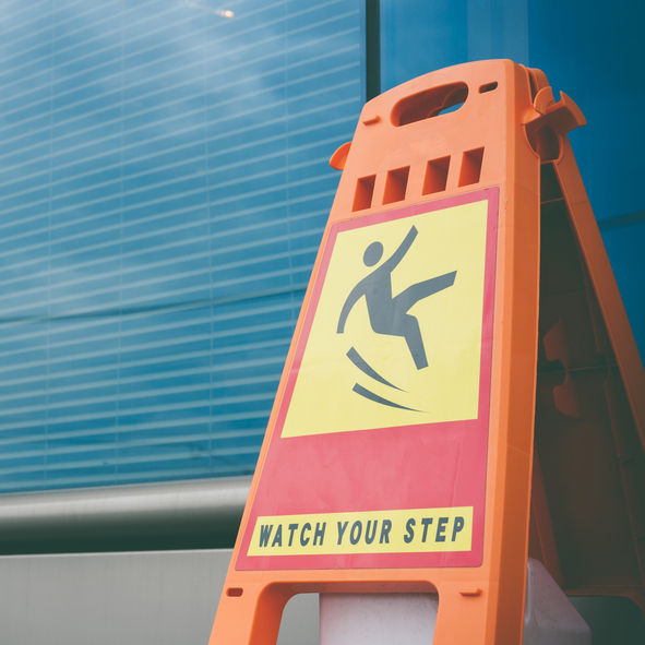 Orange caution sign with a slipping figure icon and "WATCH YOUR STEP" text, set against a blue glass building background.