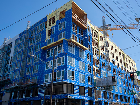 Building under construction covered in blue wrapping. Cranes and scaffolding are visible. Street signs and traffic lights in foreground.